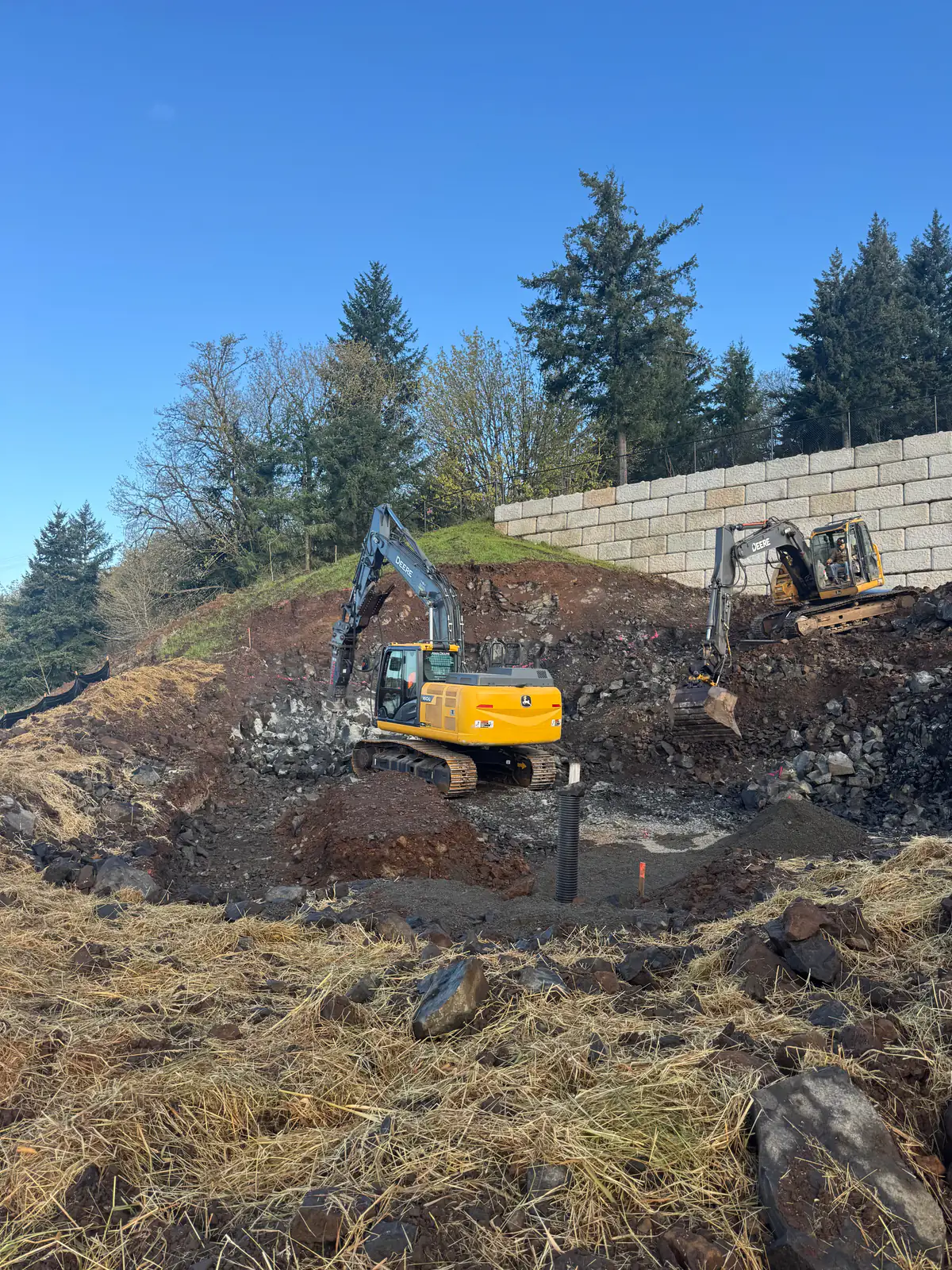 Two excavators working a rocky hillside below a block wall